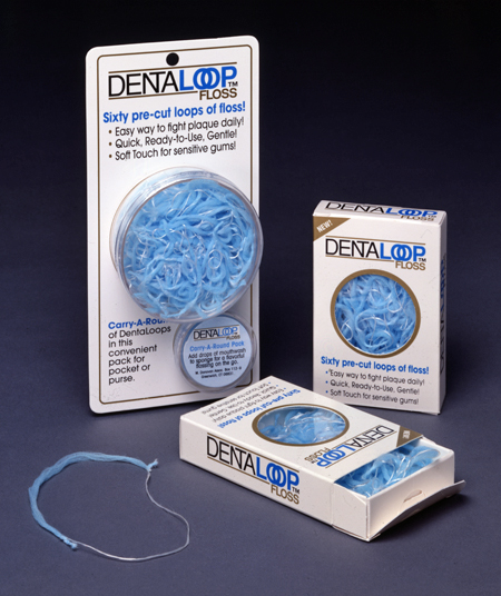 Advertising photo of 3 packages of DentaLoop floss. 2 packages stand on end and the other is flat on the table and opened to show the floss. One package holds a round container of floss, billed as a Carry-A-Round pack for pocket or purse. All packages advertise that they hold 60 pre-cut loops of the blue, circular floss, with one fluffy half and the other traditional floss.