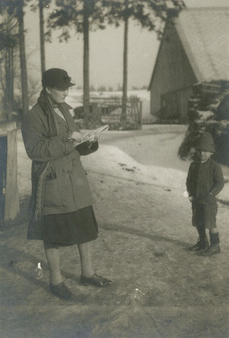 A woman outdoors, wearing a hat and coat, sketching. A young boy looks on. A log building with a steep roof is in the background.