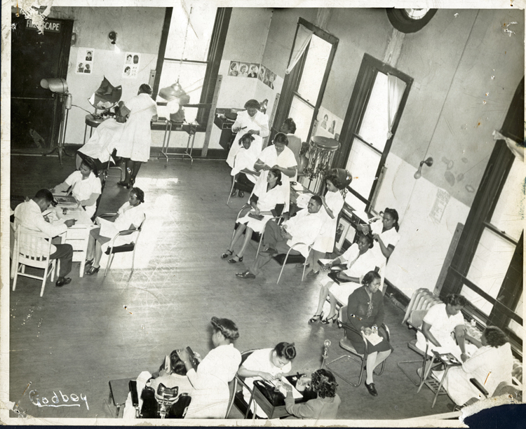 An overhead view of a beauty school classroom. 4 women, 1 man, and 1 little girl are having their hair styled. 2 women are having manicures. 2 women and 1 man are sitting at a desk, reading, and 1 woman appears to be taking notes while watching a manicurist. The room has a high ceiling and very tall windows, and photos and illustrations of hairstyles are hung on the walls.
