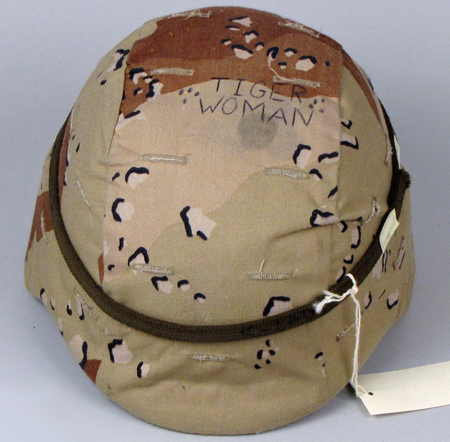 A 3/4 view from overhead of a Kevlar army helmet. The helmet is covered in tan, green, and brown camouflage-patterned cloth and the words “Tiger Woman” have been written in all capital letters on the top of the helment.