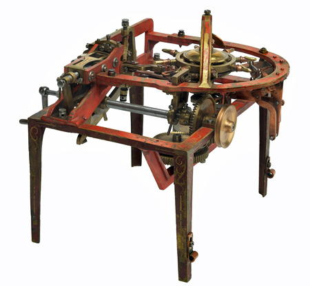 Metal model for a pin-making machine. The legs and open base are painted and ornamented in gold. The mechanism is a complicated arrangement of hand-cranked bevel gears and wire feed for extruding straight pins.