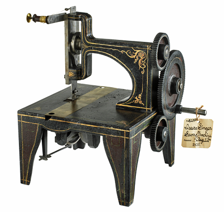 Metal model of Isaac Singer’s 1851 sewing machine, with 3 gears on the right and a sewing foot and thread feed on the left. The black metal is ornamented with gold lines and swirling patterns.