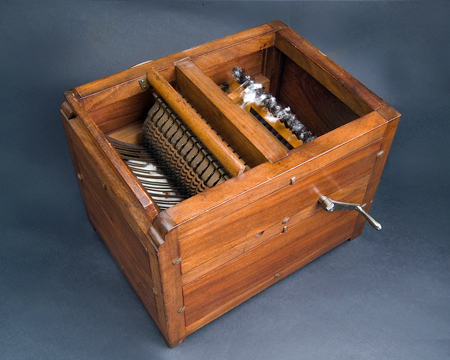 Reproduction wooden box containing several hand-cranked toothed rollers that separate cotton fibers from the seeds. Cleaned cotton fibers are collected in the base of the box.