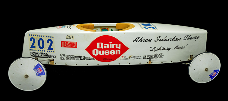 Side view of “Lightning Laura” soapbox derby car. It has a white fiberglass body and solid wheels. The car bears the words “Akron Suburban Champ Lightning Laura” and racing number 202. and is covered with various stickers, including Dairy Queen, Heath, and Continental Airlines.
