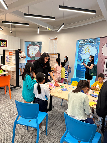 SparkLab facilitators work with a group of several young inventors at a busy and colorful activity table in front of the wind tunnel.