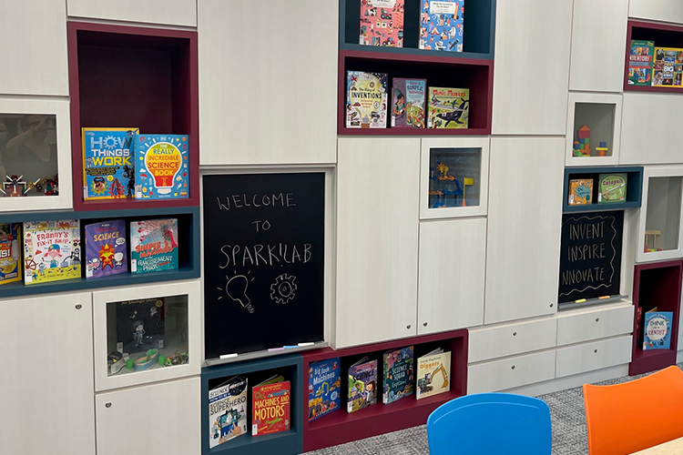 A gray bookshelf holds several books for young inventors with their covers displayed. A chalkboard in the center reads, "Welcome to Spark Lab."