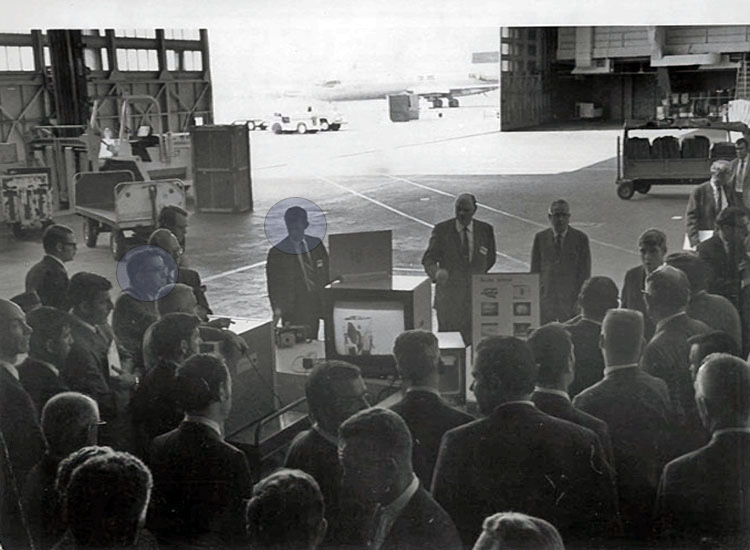 Group of men in suits inside an airplane hangar watching demonstration