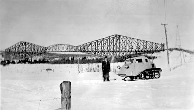 Joseph-Armand Bombardier standing outside, on snow-covered ground, wearing a topcoat and hat, and standing next to his 1937 B7 snowmobile. A truss bridge is visible in the background.