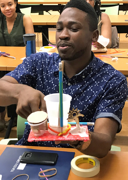 A man holds up his team’s invention, made with pencils and plastic cups. He is seated at a table and wears a dark blue-and-white patterned shirt.