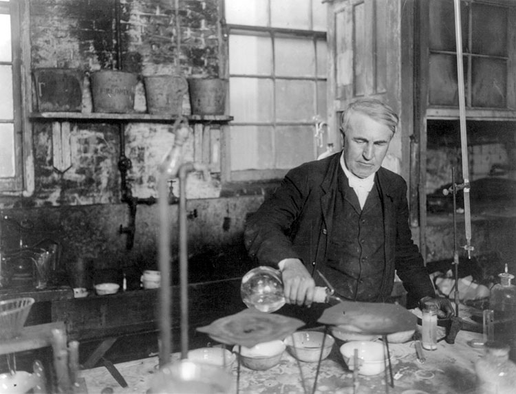 Thomas Edison pouring a liquid into a vessel on a messy lab bench in his chemistry lab