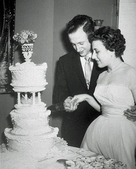 Jerome and Dorothy Lemelson at their wedding. They jointly hold the knife and are beginning to cut the wedding cake. Jerry has his arm around Dorothy’s waist.