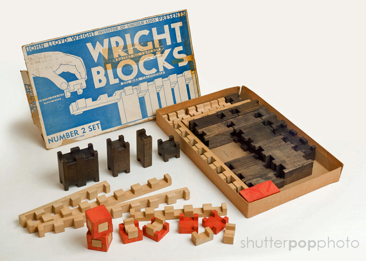 An open box of Wright Blocks. The blue-and-white box cover reads “John Lloyd Wright, Inventor of Lincoln Logs, Presents Wright Blocks, Number 2 Set. Some brown, tan, and red notched blocks are still in the box while others are arranged on the table.