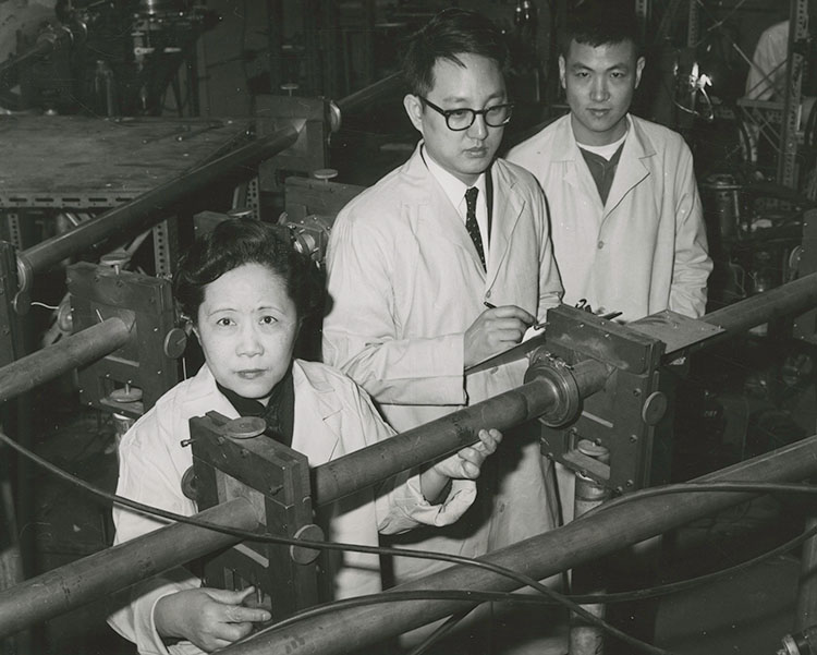 Chien-Shiung Wu with 2 other men, all in lab coats, standing behind part of a Van de Graaff accelerator.