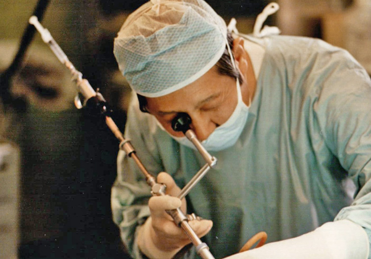 Close-up of Dr. InBae Yoon wearing a surgical gown, cap, and gloves, using a laparoscopic surgical tool. The patient is not visible in the photo.
