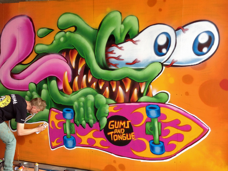 A graffiti artist is bent over, holding a spray paint can, finishing his colorful artwork that looks like a green slime monster with sharp teeth and bloodshot, bugged-out eyes, with its tongue flapping in the wind. The monster holds a skateboard reading, “Gums and Tongue.”