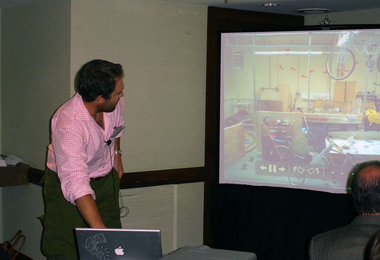 A man in a pink shirt is looking at the screen on which he is projecting a Powerpoint.