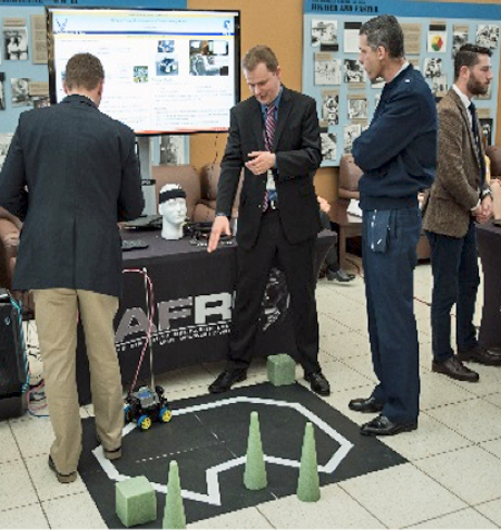 In a trade show setting, a small robot navigates an obstacle course on the floor while one man explains its movement to another man. Other men look at other displays.