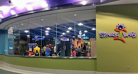 A lime green wall with the Spark!Lab logo on the right side. To the left of the wall with the logo is a long, curved wall of clear glass windows to see inside the Spark!Lab space. 