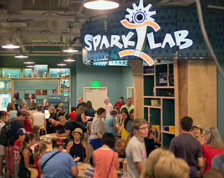 A view of the Sparklab interior space from the front of the room looking towards the back of the room. On the left and center of the picture, in the foreground is a curved table with many people standing around and some sitting on stools. In the distance 