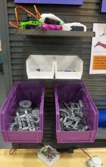close up view of two large purple plastic bins, two small white bins and one gray shelf connected to a gray wall, at the end of the activity table. In the bins are Lego pieces, some wire, and controllers.