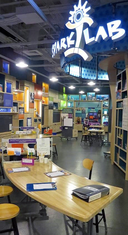 View of the Sparklab interior space from the front of the room. In the left foreground is a curved table with clipboards. In the distance is a wall with colorful display boxes, a wooden shelf unit, and the lighted sparklab sign overhead.