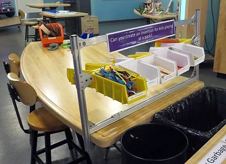 A curved, light wood table and two stools. Two vertical posts and a connecting horizontal plastic bar attached to the table hold white and yellow plastic bins filled with craft supplies in them.