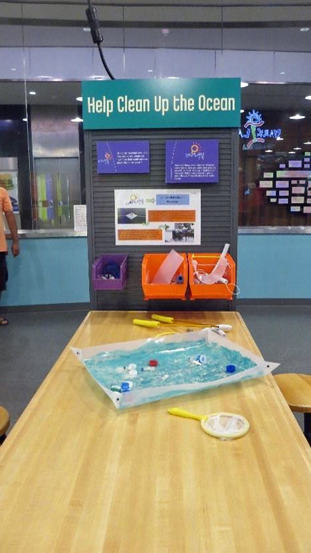A Spark!Lab activity station. A picture of an activity station, taken from the front view. In the middle of the picture is a light wooden table. teal title panel says Help Clean Up the Ocean.