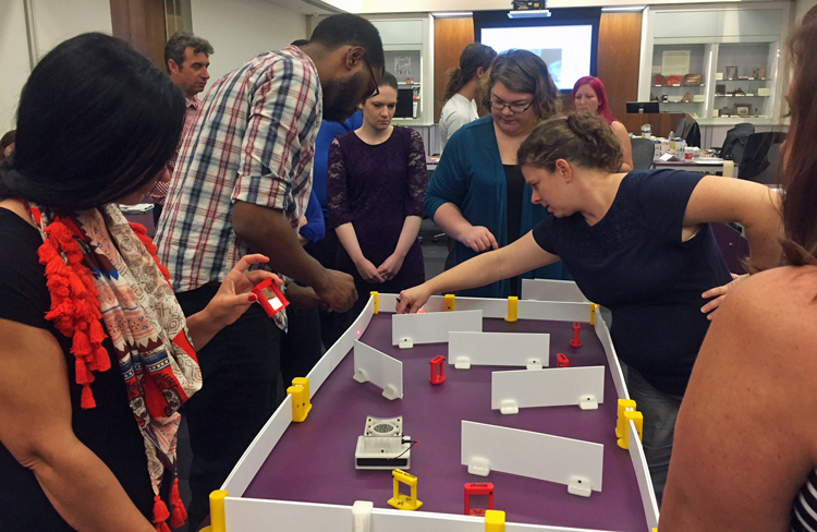 10 conference participants work on the Invent a Laser Maze activity, which sits on a table. There are fixed boards around the 4 sides of the activity, and 5 movable barriers and multiple movable targets for the laser.