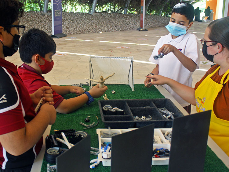 3 boys work with a SparkLab facilitator on a soccer activity; one holds a small flexible wooden mannequin in position to kick a goal.