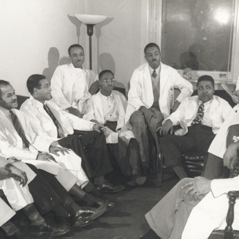 Drew smiling and addressing 9 Black medical residents seated in chairs, all wearing lab coats