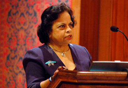 A woman speaking at a podium. She is wearing a purple suit, a silver necklace, and a large brooch of a butterfly or moth.