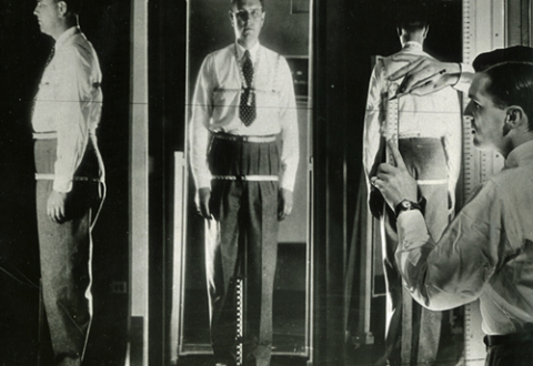 A projection of 3 views of a male customer’s reflection in mirrors, from the side, front, and back. He is wearing a tape measure harness that goes around his neck, arm (at the shoulder), chest, waist, and hips. 