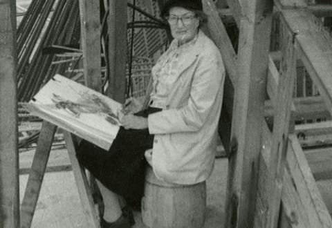 A middle-aged woman sitting on a barrel next to an outside staircase. She is holding a sketchbook.