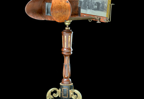 Three-quarters profile view of a free-standing stereoscope. It has curved ornamental metal legs and a carved upright support. A stereograph is in place in the holder.