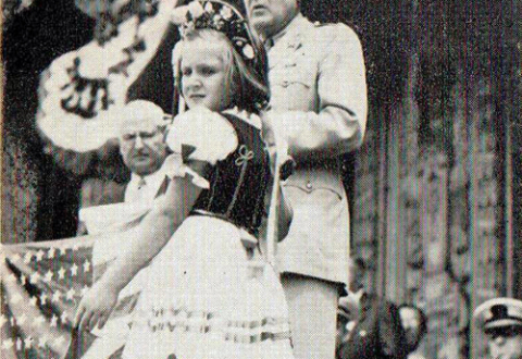 A young girl in traditional Hungarian dress looks at the camera over her shoulder while Charles Eisler at a podium introduces General Herbert Hargreaves during Eisler ambulance donation ceremony, Newark, New Jersey, 1943