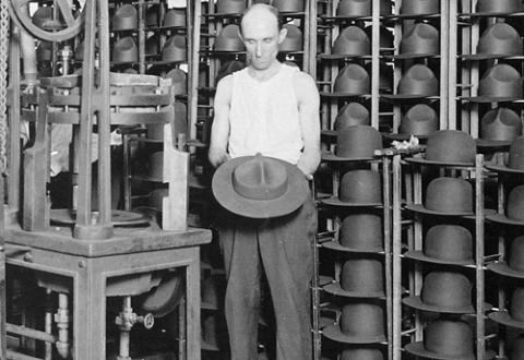 Worker standing next to a machine in the Stetson hat factory in Philadelphia, holding a hat and surrounded by racks of hats, around 1917