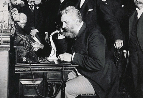 Alexander Graham Bell seated at table, speaking into telephone while a group of men watch.