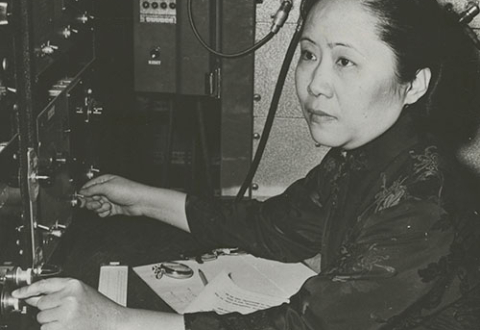 Chien-Shiung Wu in profile, sitting at a desk with a panel of knobs and dials in front of her.