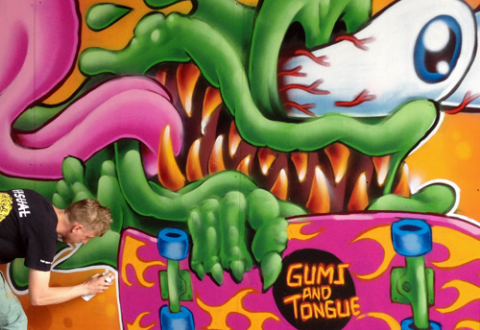 A graffiti artist is bent over, holding a spray paint can, finishing his colorful artwork that looks like a green slime monster with sharp teeth and bloodshot, bugged-out eyes, with its tongue flapping in the wind. The monster holds a skateboard reading, “Gums and Tongue.”