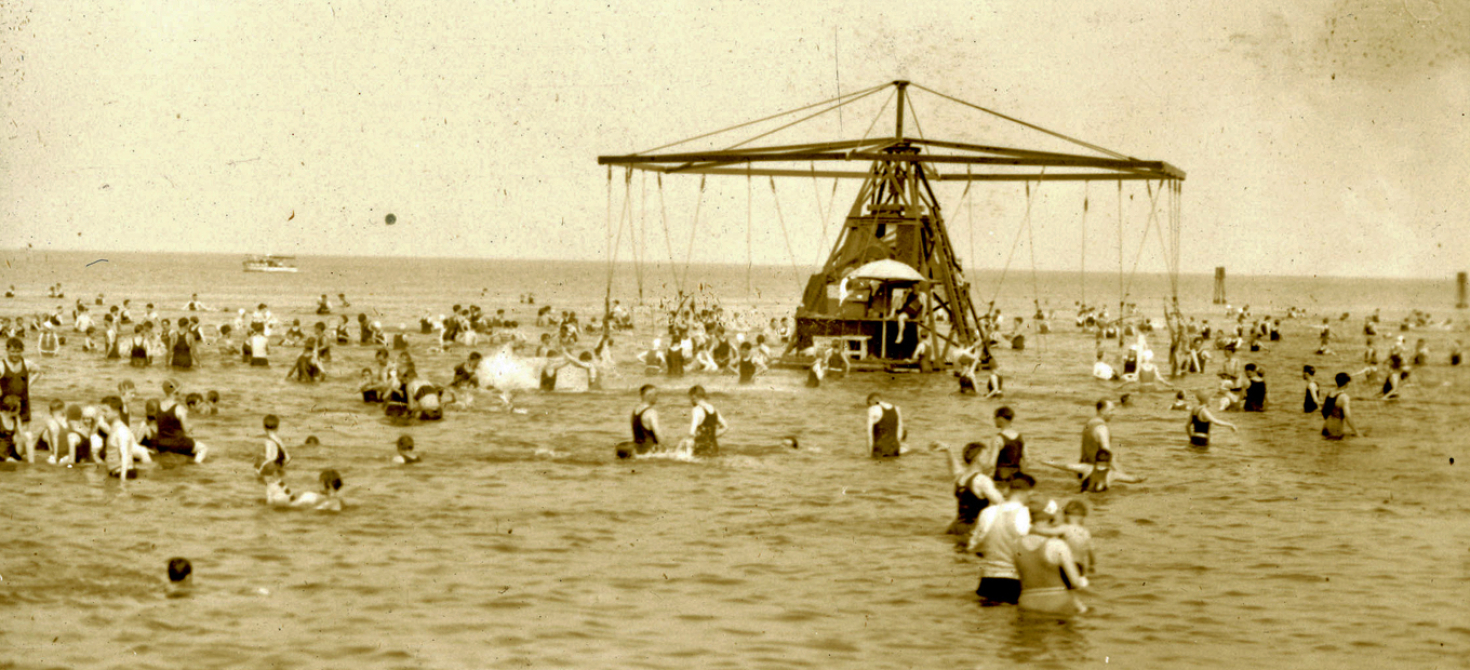 Dozens of people wading in the ocean, some of whom are riding the sea swing.