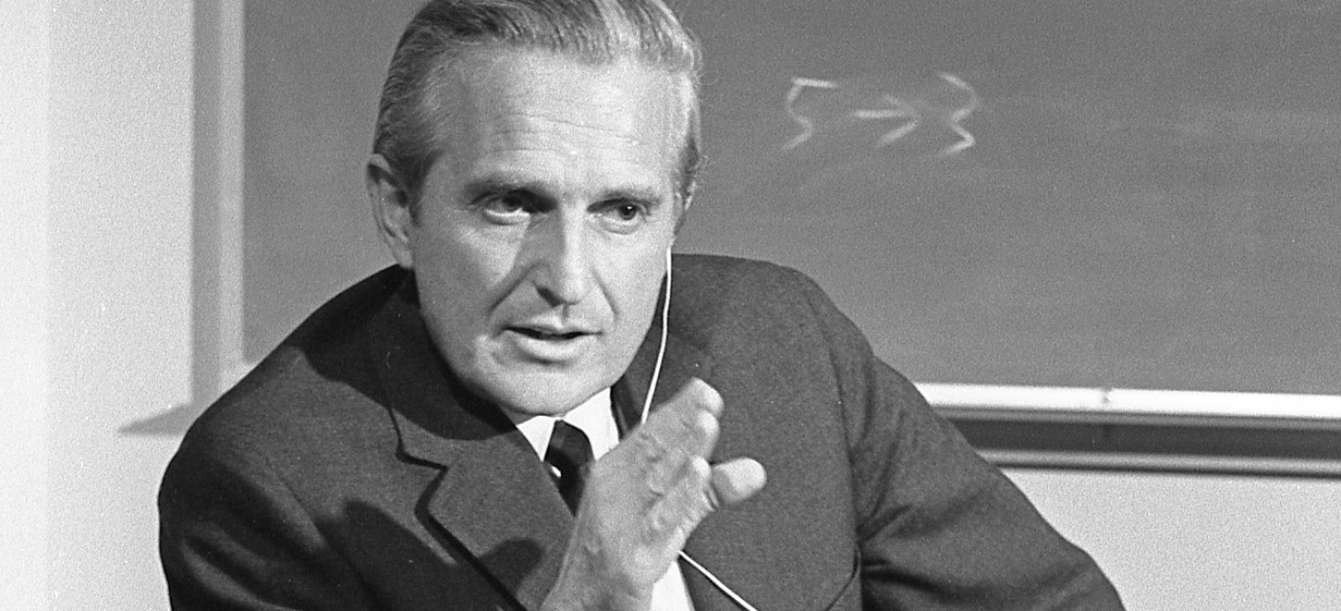 Douglas Engelbart in October 1968. He wears a jacket and tie and has a wired earpiece in his left ear. He is gesturing with his right hand and looking at someone off camera to his left. He is in front of a blackboard with the numbers 5 and 3 written on it; an arrow points from the 5 to the 3.