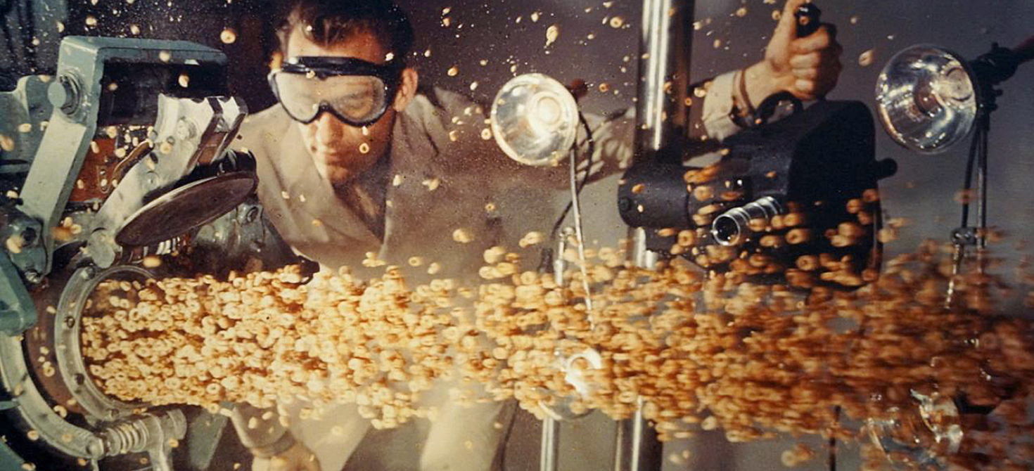 A man wearing a lab coat filming the puffed grain as it emerges from the puffing gun, undated