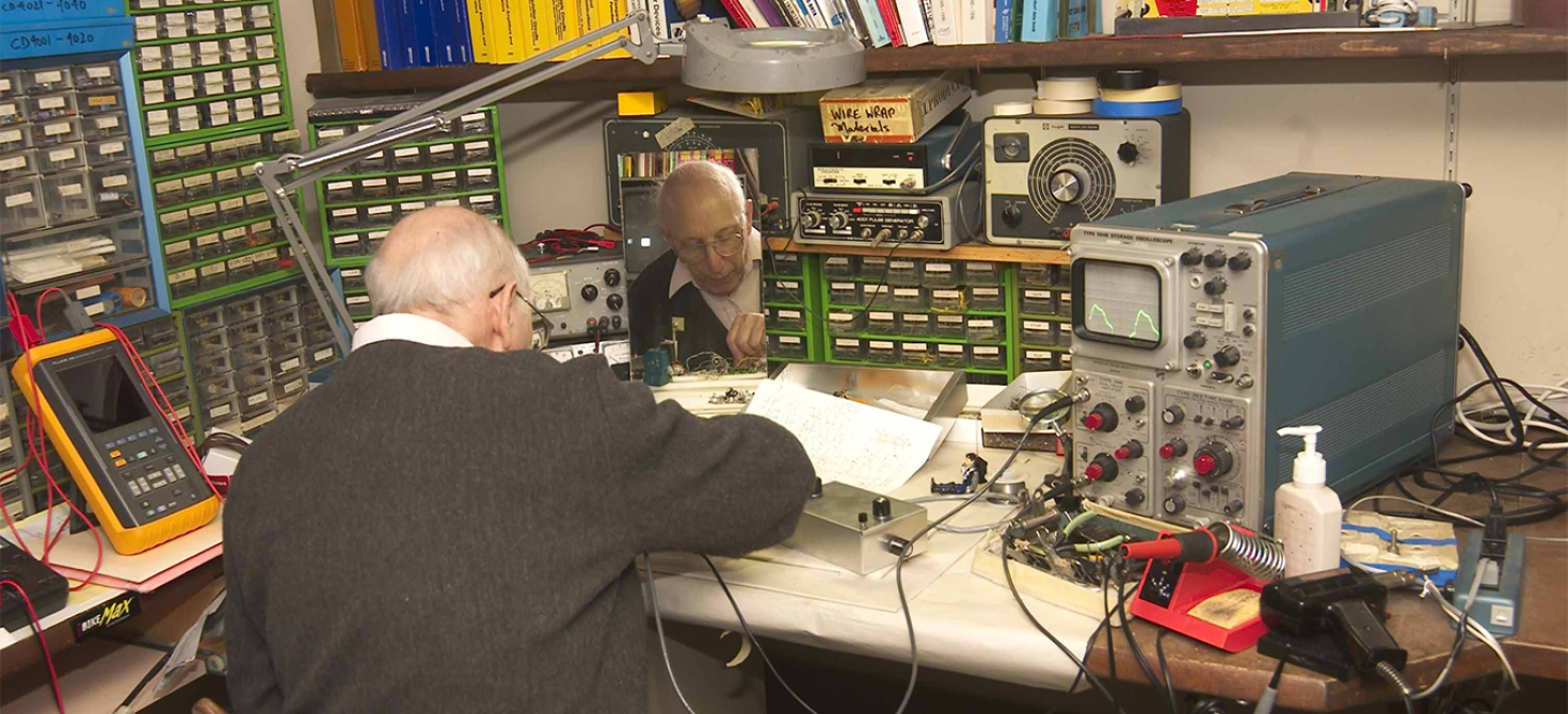 Ralph Baer sitting at his work table, with his back to the camera, working on something.