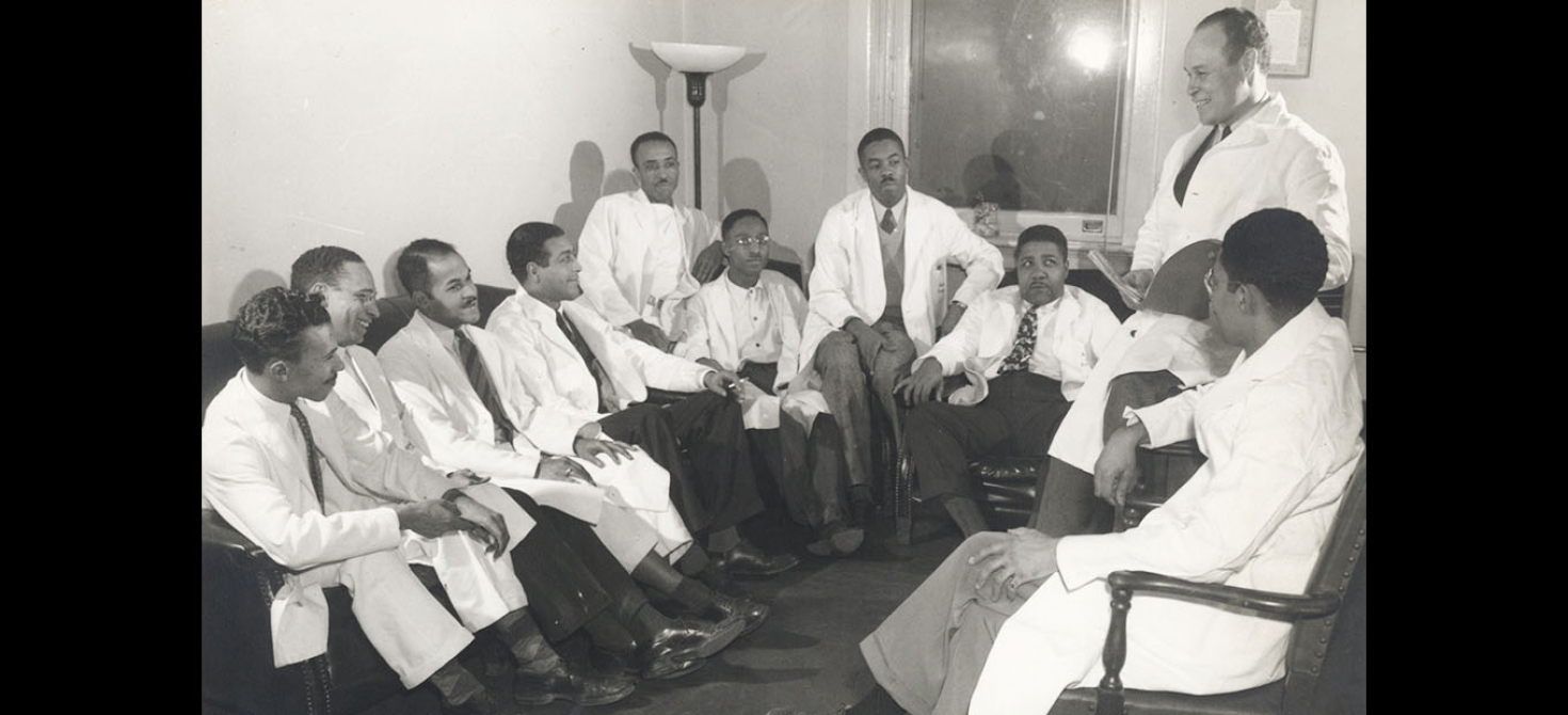 Drew smiling and addressing 9 Black medical residents seated in chairs, all wearing lab coats