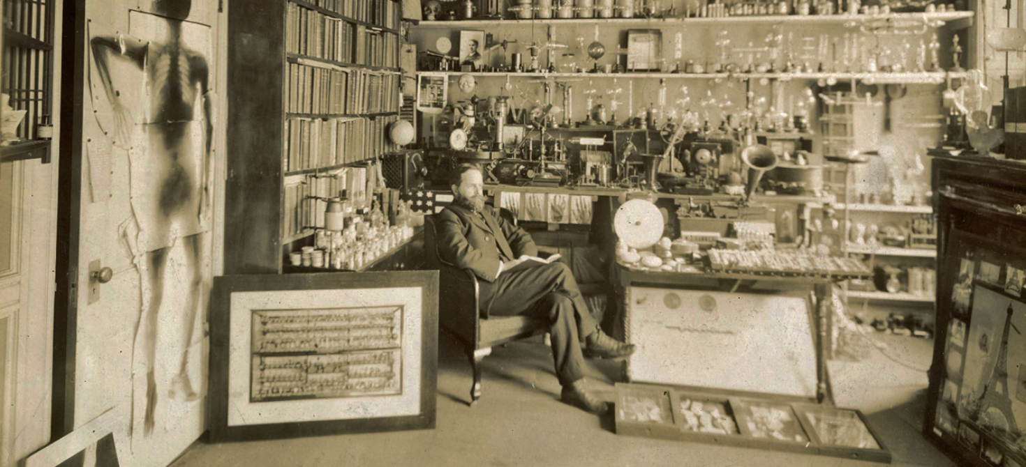 William Hammer seated in a room cluttered with scientific and experimental apparatus, including phonographs and Leyden jars