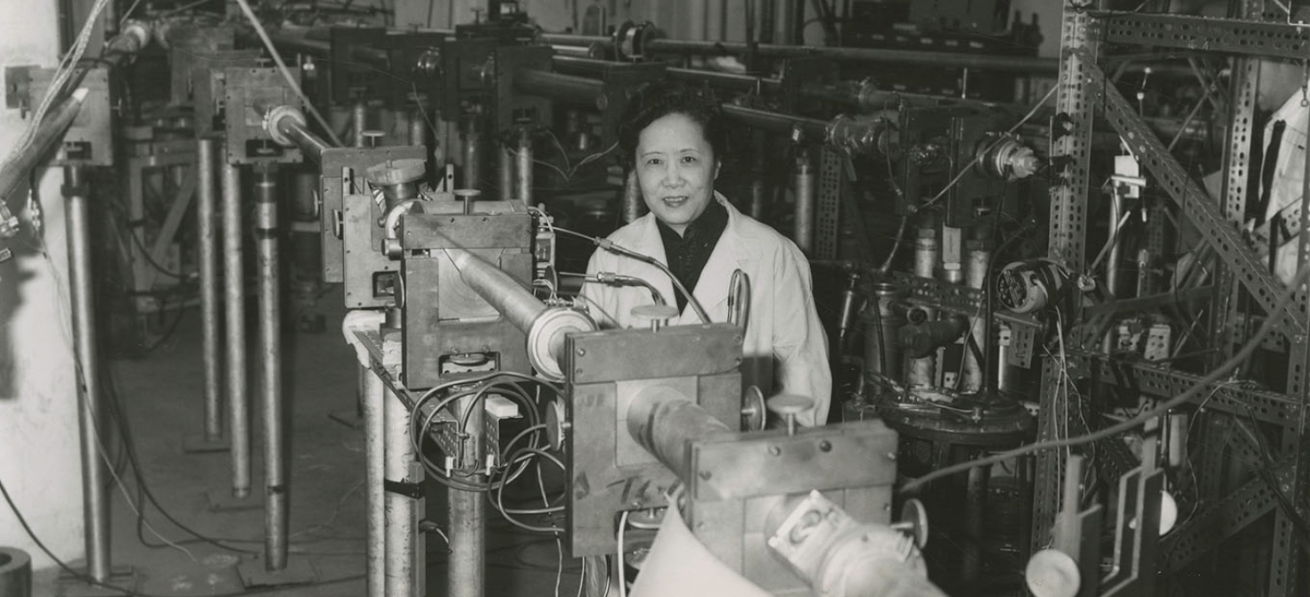 Chien-Shiung Wu, smiling at the camera and wearing a lab coat, posing among unidentified apparatus