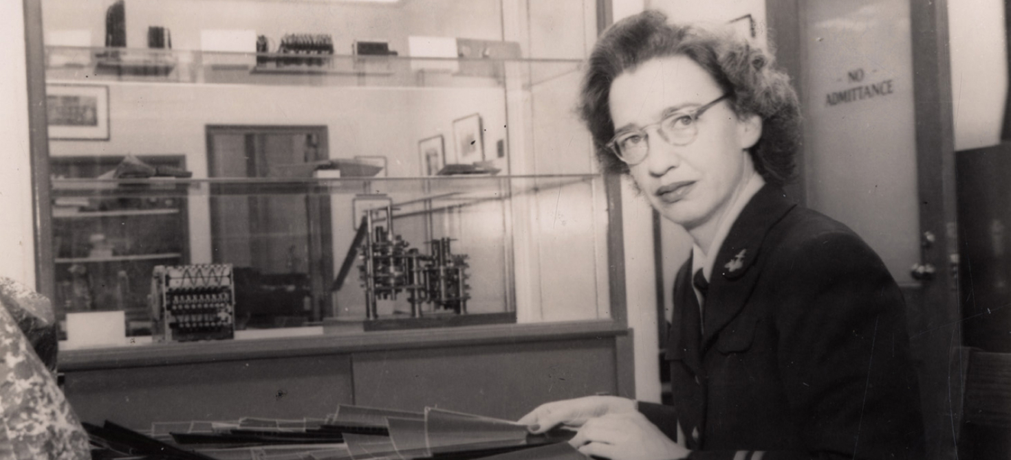 Lt. Grace Hopper at her desk in the Computation Laboratory. Glass shelves with early calculating machines seen in background, 1947.