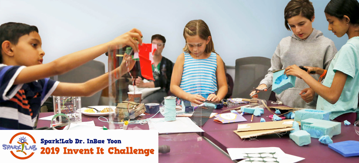 A young boy at left, a young girl in the center, and two pre-teen girls working together at right are at a table covered with craft materials. Each child is focused on his or her creative work.