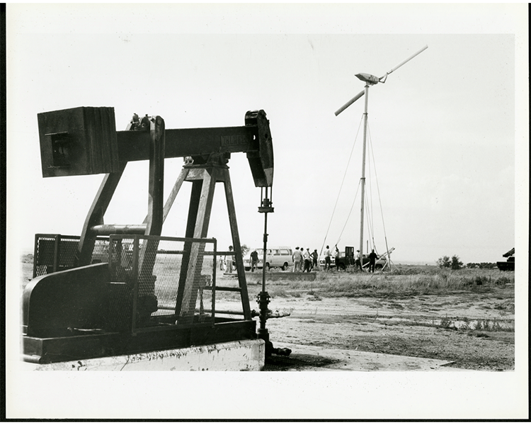 Black and white photo of a wind turbine next to an oil derrick, circa 1977-1983.