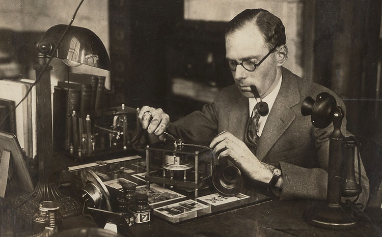 Charles Adler, in suit and tie, working on an unidentified device at his desk